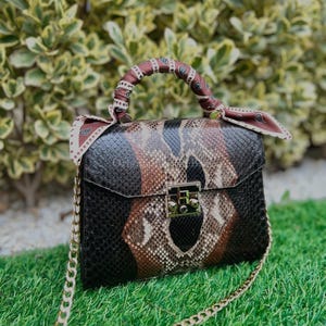 May include: A snakeskin-patterned handbag in black, brown, and tan, with a gold chain strap. The bag has a rectangular shape with a flap closure and a decorative scarf around the handle. It rests on green grass.