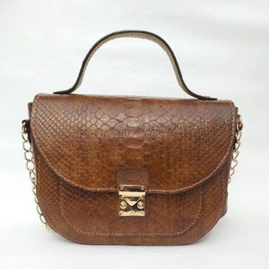 Puede incluir: Un bolso de mano de cuero marrón con un estampado de piel de serpiente. El bolso tiene forma curva, un asa superior y una correa de cadena dorada. La solapa delantera presenta un cierre dorado.