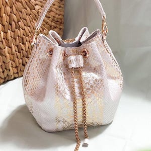 Könnte beinhalten: Eine weiße und goldene Bucket Bag mit Schlangenmuster. Die Tasche hat einen Henkel und einen Kordelzugverschluss aus goldener Kette. Die Tasche ist aus weichem Material gefertigt und hat goldfarbene Beschläge.