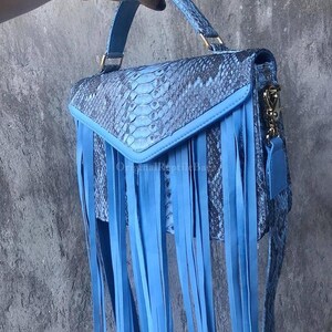 Könnte beinhalten: Eine blaue Handtasche mit Schlangenmuster und langen blauen Fransen. Die Tasche hat einen oberen Griff und einen Schultergurt. Die Tasche wird von einer Hand gehalten.