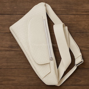 Könnte beinhalten: Cremefarbene Umhängetasche mit einem strukturierten, schuppenartigen Muster. Die Tasche hat einen langen, verstellbaren Riemen, eine Reißverschlusstasche und eine Klappentasche. Die Tasche ist aus weichem Material und hat einen silbernen Reißverschluss.