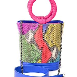 May include: A multi-coloured patterned handbag with a pink handle and a blue shoulder strap. The bag features a vibrant mix of colours, including red, yellow, green, and purple, in a snakeskin-like design. The handle is a circular design.