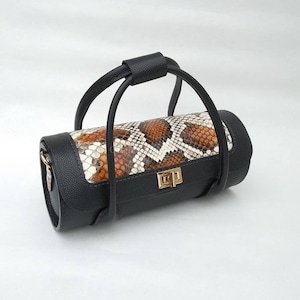Könnte beinhalten: Zylindrische Handtasche mit Schlangenmuster in Braun-, Beige- und Weißtönen. Die Tasche hat schwarze Lederakzente, einschließlich des oberen Griffs und der Seitenteile. Ein goldfarbener Verschluss sichert die Tasche.