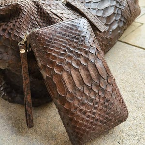 Könnte beinhalten: Eine braune Ledergeldbörse mit Handschlaufe und eine passende braune Ledertasche. Die Geldbörse und die Tasche haben eine schuppenartige Textur. Die Geldbörse ist rechteckig und hat einen Reißverschluss. Die Tasche hat einen Reißverschluss.