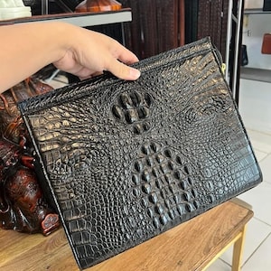 Könnte beinhalten: Schwarze Leder-Clutch mit Krokodilhaut-Textur. Die rechteckige Tasche hat einen Reißverschluss und ein detailliertes geprägtes Muster. Die Tasche wird von einer Hand gehalten, die die Textur und das Design hervorhebt.