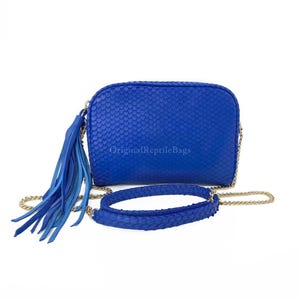 Könnte beinhalten: Eine leuchtend blaue Umhängetasche mit einem strukturierten, schuppenartigen Muster. Die Tasche hat einen langen, goldenen Kettenriemen und einen passenden blauen Riemen. Eine lange Quaste hängt an der Seite. Der Text "Original Reptile Bags" ist sichtbar.