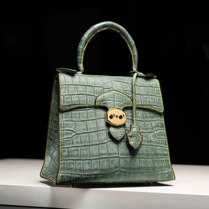 Puede incluir: Bolso de mano verde salvia con textura de piel de cocodrilo. El bolso tiene un asa superior, un cierre de solapa con un cierre dorado y herrajes dorados. Las costuras son de un color amarillo-verdoso contrastante.