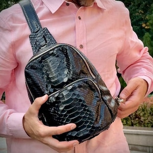 Könnte beinhalten: Eine schwarze, strukturierte Leder-Umhängetasche mit Reißverschluss. Die Tasche hat einen Schultergurt und wird über der Brust getragen. Die Oberfläche der Tasche hat ein schuppenartiges Muster.