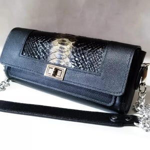 Puede incluir: Un bolso de mano negro y gris con una correa de cadena plateada y una correa de hombro de cuero negro. El bolso presenta un diseño texturizado con un panel de piel de serpiente y un cierre de broche plateado. El bolso mide aproximadamente 25 cm de largo.