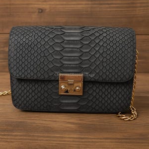May include: A black, rectangular handbag with a textured snakeskin pattern. The bag features a gold-colored clasp and a gold chain strap. The bag is made of a dark material and has a flap closure.