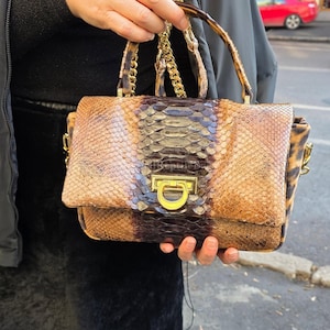 Puede incluir: Un bolso rectangular con un estampado de piel de serpiente en tonos marrón, beige y morado. El bolso presenta un cierre y una correa de cadena dorados. El asa tiene un diseño de carey. El bolso lo sostiene una persona.