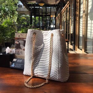 Könnte beinhalten: Eine weiße, strukturierte Handtasche mit goldenen Kettenriemen. Die Tasche hat eine Eimerform und ein Schlangenmuster. Die Tasche steht auf einem Holztisch. Der Hintergrund umfasst Grün und ein Gebäude.