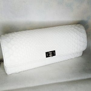 Könnte beinhalten: Eine weiße Clutch mit einem strukturierten, schlangenlederartigen Muster. Die rechteckige Tasche hat einen Klappverschluss mit einem silberfarbenen Verschluss. Eine zarte Silberkette ist an der Seite sichtbar. Ein neutrales Accessoire.