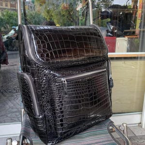 Op de afbeelding: Een zwarte rugzak met een glanzende krokodillenleertextuur, voorzien van een grote klep, een voorvak met rits en zijvakken met rits. De tas is afgewerkt met zilverkleurige metalen ritsen en een subtiel in reliëf aangebracht logo op het voorvak.