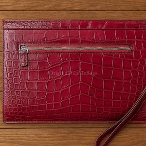 Könnte beinhalten: Eine rote Leder-Clutch mit Krokodilhaut-Textur. Die rechteckige Tasche hat einen silbernen Reißverschluss und eine Handschlaufe. Die Tasche liegt auf einer Holzoberfläche.