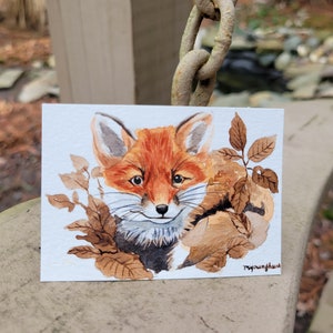 Op de afbeelding: Waterverf schilderij van een rode vos met bruine vacht die tevoorschijn komt achter bruine bladeren. De vos kijkt rechtstreeks naar de kijker met een lieve uitdrukking. De handtekening van de kunstenaar, "myounghwa", staat in de rechter benedenhoek.