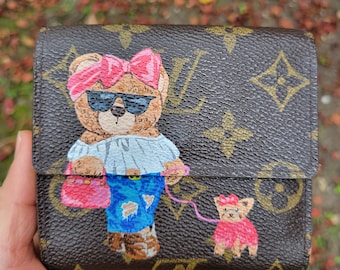 May include: Brown and tan wallet with a painted bear design. The bear wears sunglasses, a pink bow, and a light blue shirt. It holds a pink purse and walks a small dog. The wallet has a brown monogram pattern.