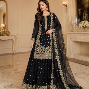 May include: A black long-sleeved tunic and matching skirt set featuring intricate gold embroidery along the hem, neckline, and bodice. The ensemble includes a sheer black dupatta with a decorative gold embroidered border and scattered gold accents.