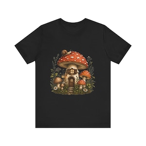 Könnte beinhalten: Ein schwarzes Kurzarm-T-Shirt mit einem zentralen Aufdruck eines großen Pilzhauses mit rotem Hut, das eine Tür und Fenster besitzt, umgeben von kleineren Pilzen, grünem Blattwerk, weißen Blumen und einer Schnecke auf dem Hauptpilz.