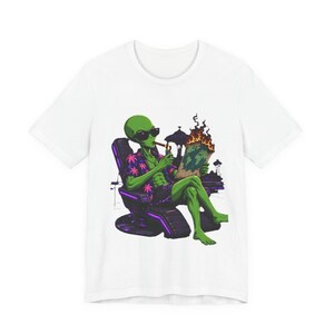 Könnte beinhalten: Ein weißes T-Shirt mit einer Grafik eines grünen Außerirdischen, der eine Sonnenbrille und ein Blumenhemd trägt, in einem Sessel sitzt, raucht und eine brennende Weltkarte hält. Das Design verwendet leuchtende violette und orangefarbene Akzente.