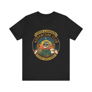 Könnte beinhalten: Ein schwarzes T-Shirt mit einem kreisförmigen Aufdruck und dem Text COFFEE & SUNSHINE ADVENTURE CLUB EXPLORE CREATE INSPIRE. Das Motiv zeigt eine Kaffeetasse mit Sonnenbrille und Schnurrbart, Palmen, einen Sonnenuntergang und Hände mit Malutensilien.