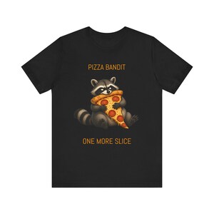 Könnte beinhalten: Ein schwarzes T-Shirt mit einer Grafik eines Waschbären, der ein Stück Pepperoni-Pizza isst. Der Text PIZZA BANDIT ist in Orange über dem Waschbären gedruckt, und der Text ONE MORE SLICE ist in Orange unter dem Waschbären gedruckt.