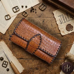 May include: A tan rectangular wallet with a textured crocodile skin pattern, featuring dark brown stitched edges and a central strap closure. The wallet is positioned among various metal buckles, tools, and pieces of paper on a wooden surface.