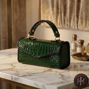 Peut inclure: Un sac à main rectangulaire d'un vert émeraude profond avec une texture brillante façon crocodile. Le sac présente une poignée supérieure structurée et une fermeture à rabat, toutes deux ornées de surpiqûres noires épaisses sur les bords.