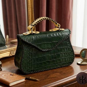 Könnte beinhalten: Eine dunkelgrüne Handtasche mit Krokodilleder-Textur und einem Klappverschluss mit schwarz vernähten Kanten. Der obere Griff ist hellgelb mit schwarzer Kreuzstichnaht und mit silberfarbenen Metallbeschlägen an der Tasche befestigt.
