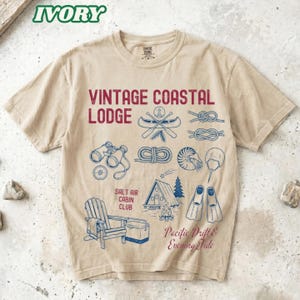 May include: An ivory short-sleeve t-shirt featuring red and blue graphic prints. The design includes the text "VINTAGE COASTAL LODGE," "SALT AIR CABIN CLUB," and "Pacific Drift & Evening Tide," alongside illustrations of binoculars, a canoe, knots, a shell, a cabin, a campfire, and swim fins.