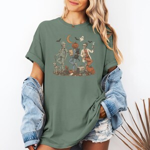 May include: A sage green t-shirt featuring a graphic illustration of three skeleton figures dressed as veterinary staff. The design includes a pumpkin-headed figure, a black cat, a dog, a ghost, bats, a crescent moon, and various medical supplies.