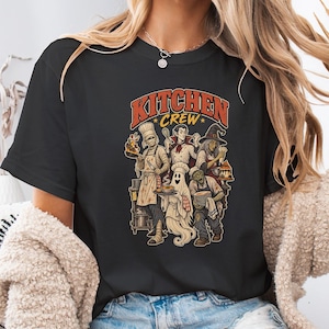 May include: A black t-shirt featuring a graphic print with the text KITCHEN CREW in orange letters. The illustration depicts a mummy, a vampire, a witch, a ghost, and a zombie dressed as chefs, holding various kitchen tools and food items.