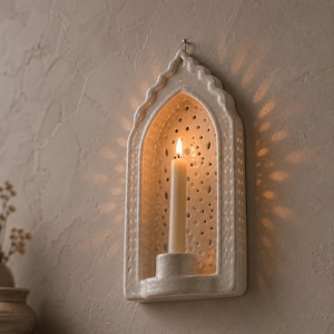 Puede incluir: Un aplique de pared de cerámica color crema con una parte superior arqueada y puntiaguda y patrones perforados intrincados. El aplique sostiene una vela cónica delgada de color blanco roto que está encendida, proyectando luz cálida.