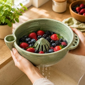 May include: A sage green ceramic berry colander bowl featuring a central integrated reamer cone, a side spout, and a handle with five small drainage holes. The bowl is filled with fresh red strawberries, blueberries, and blackberries being rinsed.