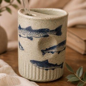 May include: A cylindrical ceramic tumbler with a speckled beige finish and a blue fish pattern. The vessel features a thumb indentation on the side, a ribbed base, and a lid with a small opening for a clear glass straw to pass through the top.