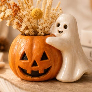 May include: A ceramic planter shaped like an orange jack-o'-lantern pumpkin with a carved face, featuring a glossy white ghost figure hugging the side. The pumpkin is filled with a bouquet of dried yellow and beige flowers and grasses.