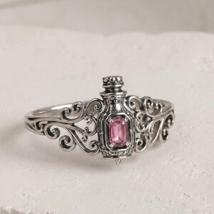 Könnte beinhalten: Ein Silberring mit einem flaschenförmigen Mittelstück, das einen rosafarbenen Edelstein im Smaragdschliff enthält. Die Ringschiene ist mit filigranen, geschwungenen Metallverzierungen versehen, die sich vom zentralen Motiv aus erstrecken.