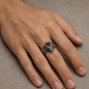 May include: A silver-toned metal ring featuring a detailed snake head design with a small skull positioned on top. The band of the ring displays an intricate, embossed pattern across its surface, providing a textured and metallic appearance.