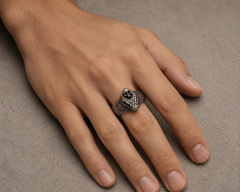 May include: A silver-toned metal ring featuring a detailed snake head design with a small skull positioned on top. The band of the ring displays an intricate, embossed pattern across its surface, providing a textured and metallic appearance.
