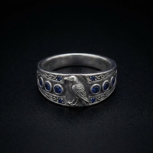 May include: A silver-toned metal ring featuring a central embossed raven figure surrounded by a series of small, round, deep blue stones set into the band. The metal surface has a darkened, oxidised finish with intricate engraved knotwork patterns.