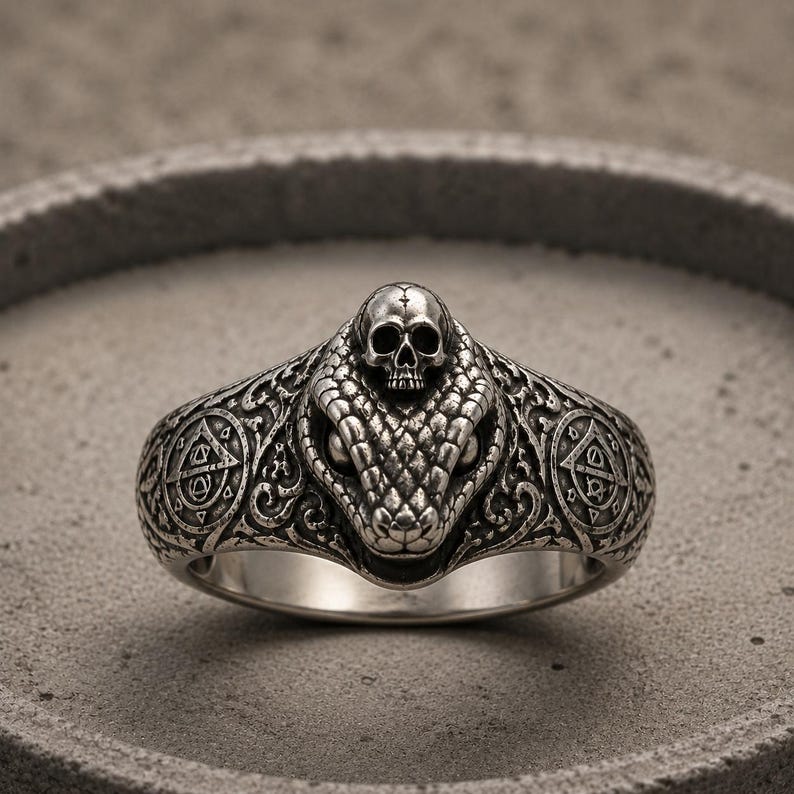 May include: A silver ring featuring a detailed snake head with a small skull resting on top. The band is decorated with intricate scrollwork and circular symbols on both sides, showcasing a dark, oxidised finish that highlights the metal textures.