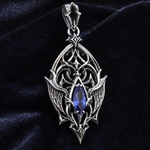 May include: A silver pendant featuring an intricate, pointed metal frame with symmetrical wing-like details and scrollwork. A single, marquise-cut, deep blue gemstone is set vertically in the centre of the design, topped by a decorative bail.