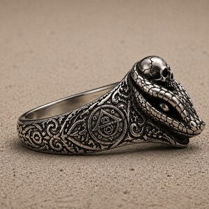 May include: A silver-toned ring featuring a detailed snake head design with a small skull resting on top. The band is decorated with intricate scrollwork and a circular emblem containing a triangle and geometric symbols in a dark, oxidised finish.