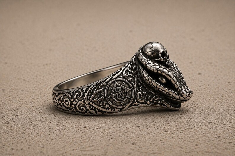May include: A silver-toned ring featuring a detailed snake head design with a small skull resting on top. The band is decorated with intricate scrollwork and a circular emblem containing a triangle and geometric symbols in a dark, oxidised finish.