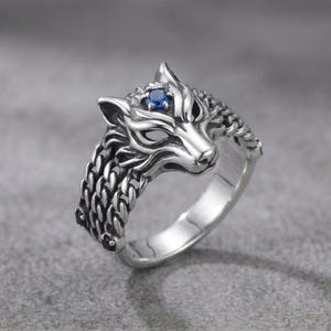 May include: A silver-colored ring featuring a wolf's head design. The wolf has a blue gemstone eye and is surrounded by a chain-like band. The ring is set against a gray background.