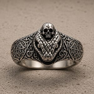 May include: A silver ring featuring a detailed snake head with a small skull resting on top. The band is decorated with intricate scrollwork and circular symbols on both sides, showcasing a dark, oxidised finish that highlights the metal textures.