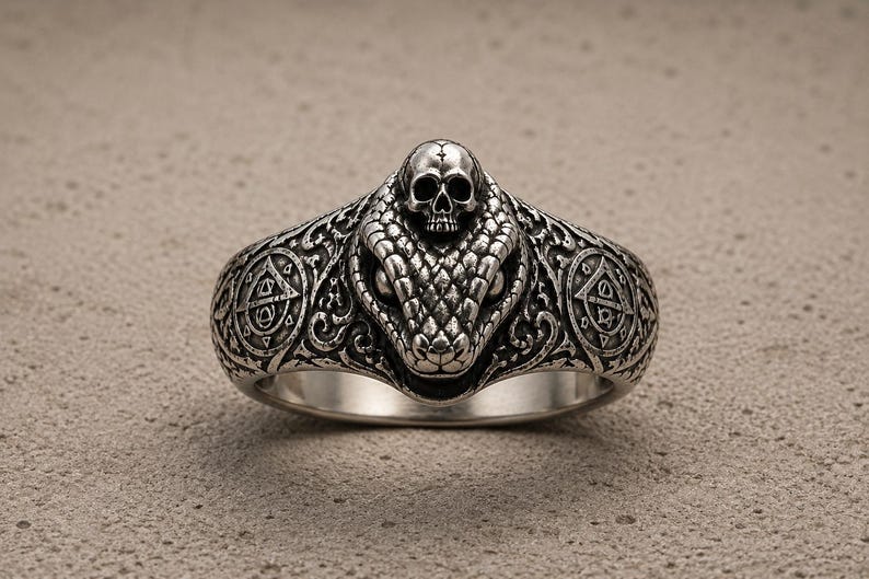 May include: A silver ring featuring a detailed snake head with a small skull resting on top. The band is decorated with intricate scrollwork and circular symbols on both sides, showcasing a dark, oxidised finish that highlights the metal textures.