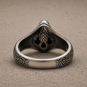 May include: A silver-toned metal ring featuring an intricate, textured relief design on the band and a central, raised ornament shaped like a stylised acorn or pinecone with dark, recessed detailing throughout the metal surface.