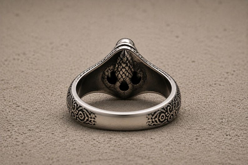 May include: A silver-toned metal ring featuring an intricate, textured relief design on the band and a central, raised ornament shaped like a stylised acorn or pinecone with dark, recessed detailing throughout the metal surface.