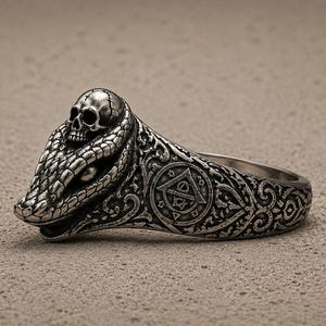 May include: A silver-toned metal ring featuring a detailed snake head with a small skull resting on top. The band is decorated with intricate scrollwork and a circular emblem containing a triangle and geometric symbols on the side.
